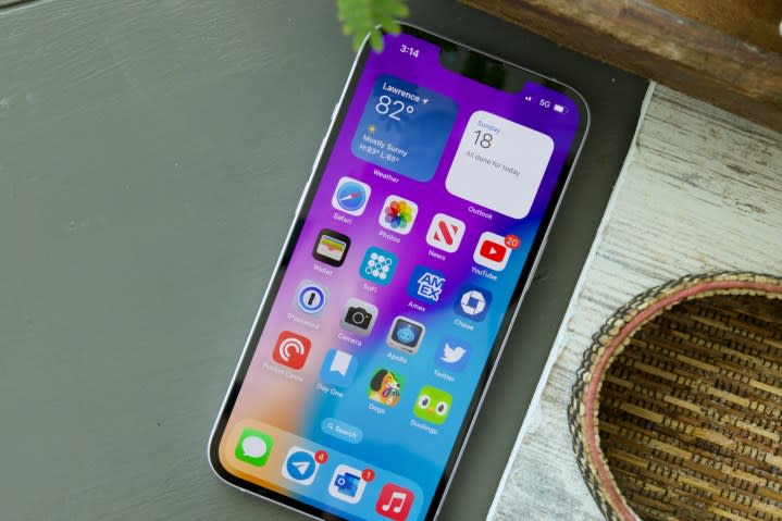 An iPhone 14 on a table. The screen is on and displays the home screen.